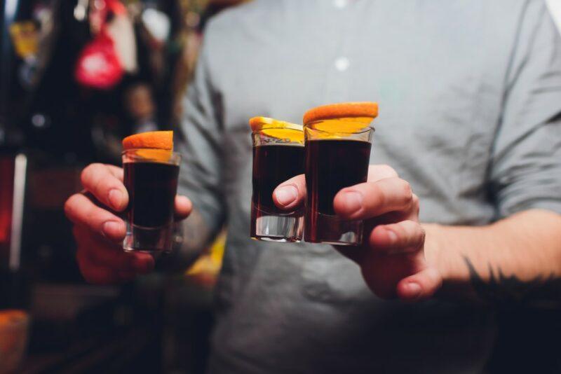 A man holding three Hell's fury shots in his hand, each garnished with a piece of orange