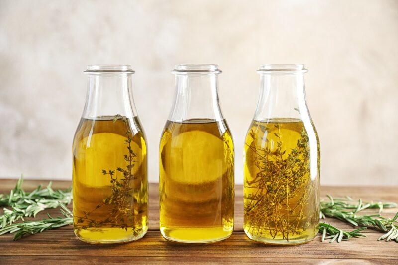 Three small glass bottles containing olive oil. The outside two are infused with herbs, while the middle one isn't. There are herbs on the table too