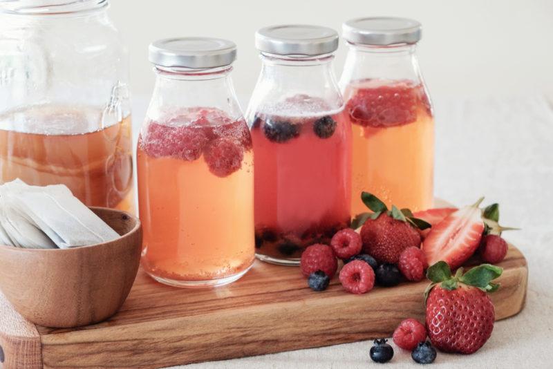 A wooden board with three bottles of kombucha, each containing a different type of fruit, there are tea bags, a bottle of plain kombucha, and various fresh fruits too