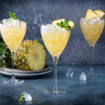 Three caribou Lou cocktails with ice on a slate table with a pineapple in the background