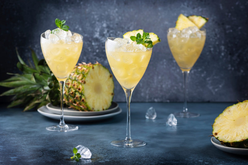 Three caribou Lou cocktails with ice on a slate table with a pineapple in the background