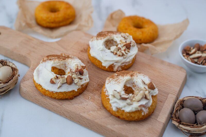 A wooden board with three carrot cake donuts with cream cheese frosting. There are some more donuts and other ingredients in the background