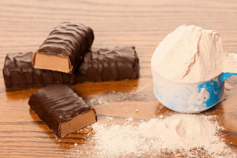 A wooden table with three chocolate covered protein bars and a blue scoop containing vanilla protein powder