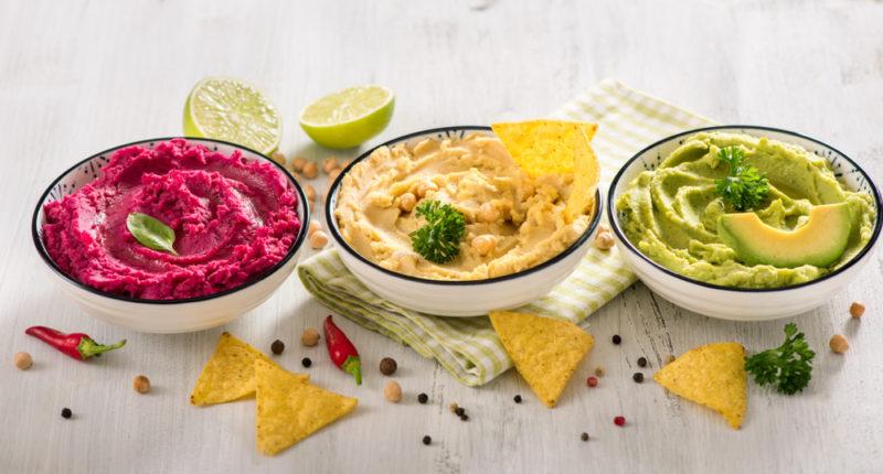 Three white bowls of vegan dip with food to dip in them scattered around