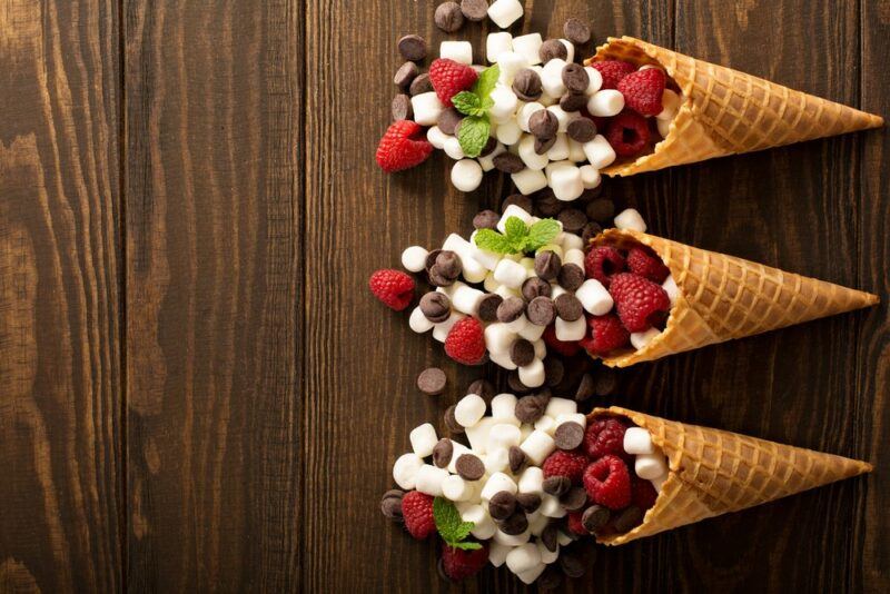 A wooden table with three dessert cones that have raspberries, chocolate, and marshmallows spilling out of them