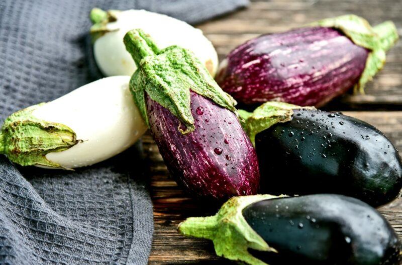 Six eggplants on a wooden table, two are purple, two are purple and white, while the remaining two are white