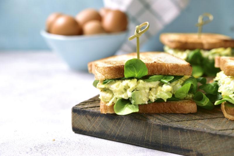 Three egg and avocado sandwiches on a wooden board with eggs in the background