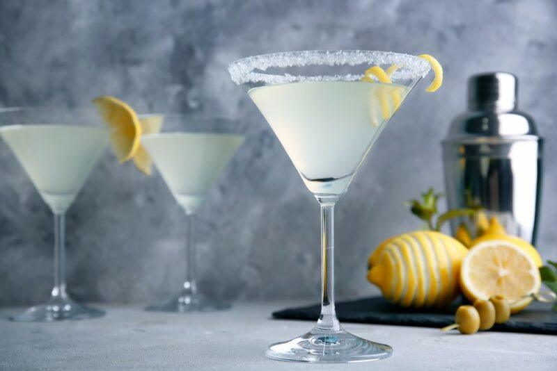 Three lemon drop martinis in glasses with sugar rims and lemon slices, next to a cocktail shaker and some whole lemons