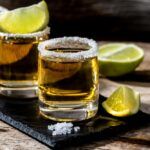 A black slate with three shots called 151 ways to die, each rimmed with salt. One has a lime wedge on top and there are limes on the table too.