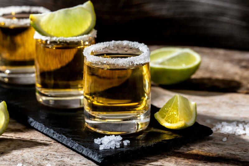 A black slate with three shots called 151 ways to die, each rimmed with salt. One has a lime wedge on top and there are limes on the table too.