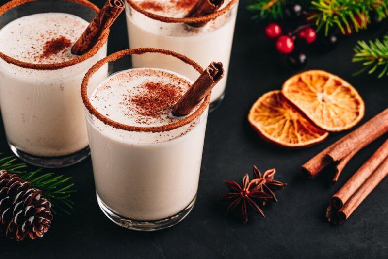 Three glasses of a coquito cocktail on a black background with cinnamon sticks as a garnish, next to slices of oranges, cranberries, star anise, and cinnamon