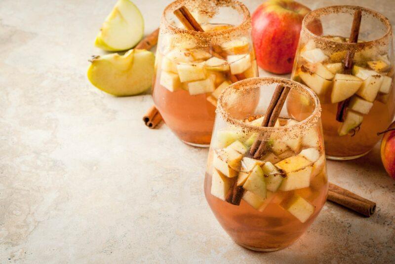 Three glasses of a fall punch cocktail on a light table with cubes of apple and cinnamon sticks as a garnish