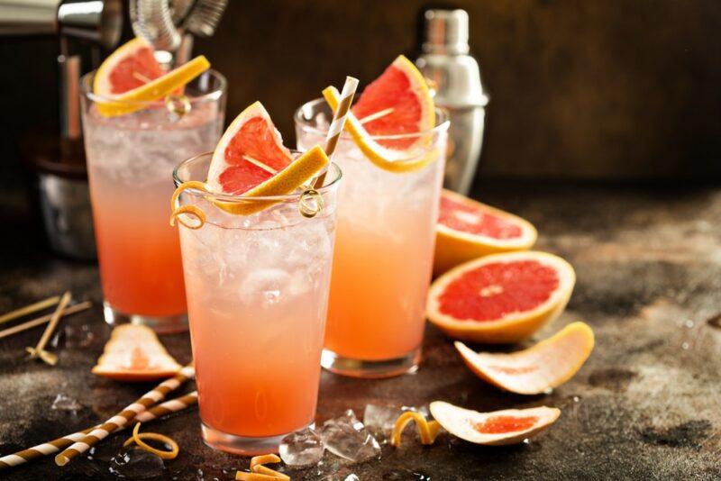 Three glasses of a fizzy dog cocktail, each garnished with grapefruit, with more pieces of grapefruit on the table