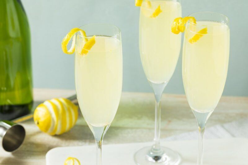 Three champagne glasses containing a lemosa cocktail and garnished with a twist of lemon, next to a bottle and a lemon on a table