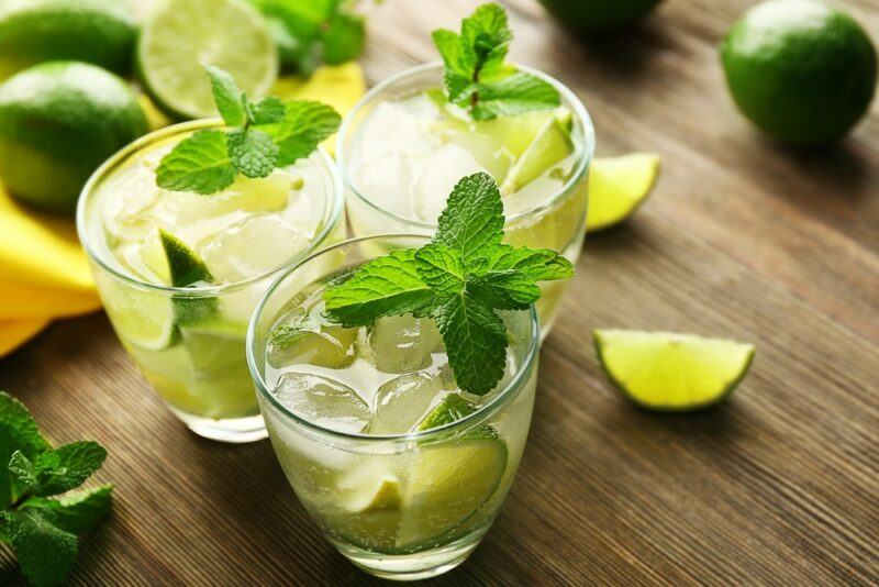 A wooden table with three glasses of a mojito cocktail, each garnished with peppermint. There are also various limes on the table