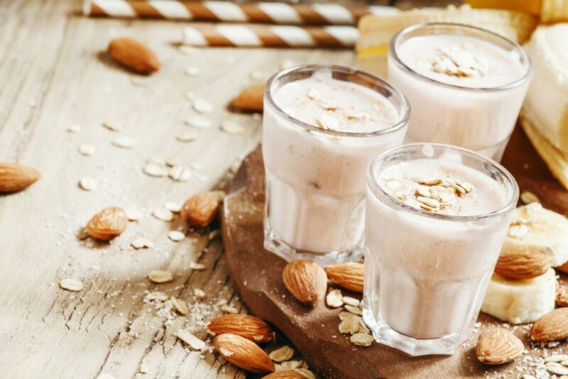 Three glasses of an almond milk smoothie on a wooden board, with straws in the background and almonds scattered around