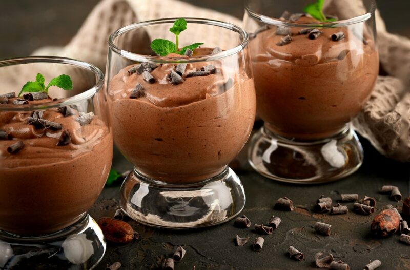 A dark table with three glasses containing chocolate mousse, with chocolate chips on top and peppermint leaves, with more chocolate chips on the table