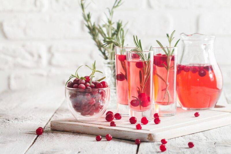 A wooden board with a glass bowl of cranberries, three glasses of cranberry juice with water, and a jug of the juice and water combination