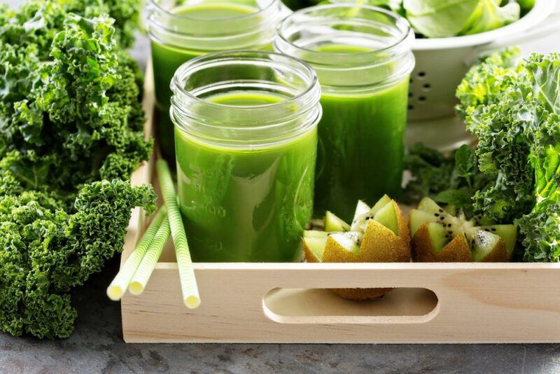 A tray with glasses of a kale cocktail next to bunches of kale and some fruit