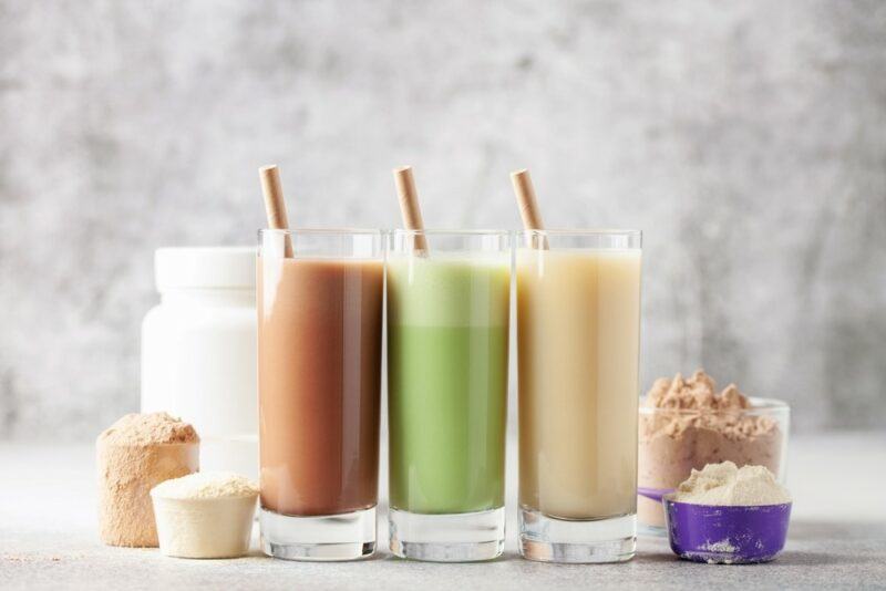 Three glasses of plant-based protein powder shakes, with scoops of the powder around them