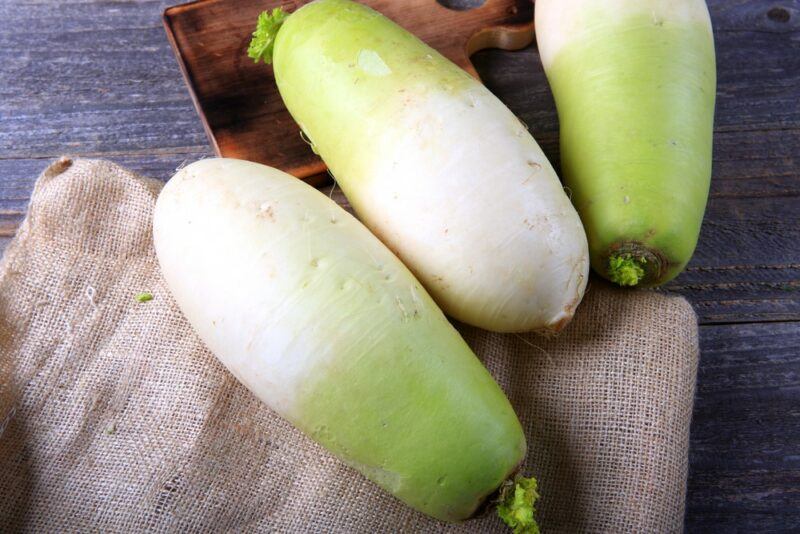 Three large Korean radishes, all with a white base and a green top. They're on a table with a cloth and a wooden board.