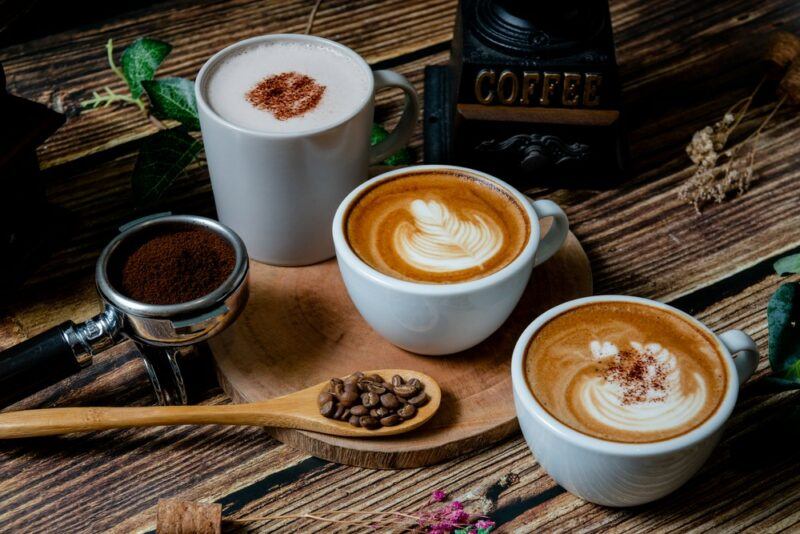 Three mugs with different types of coffee, next to an espresso shot and a spoon of coffee beans