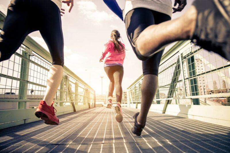 Three people running outside on a bridge. The one in front is a woman with a pink top and a long pony tail