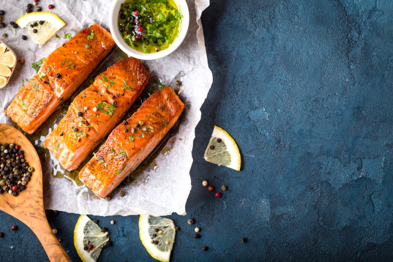 Three pieces of baked salmon on a piece of parchment paper with seasonings and lemon wedges scattered around