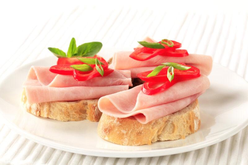 A white plate with three pieces of bread that have sliced chicken ham, tomatoes, and some greens