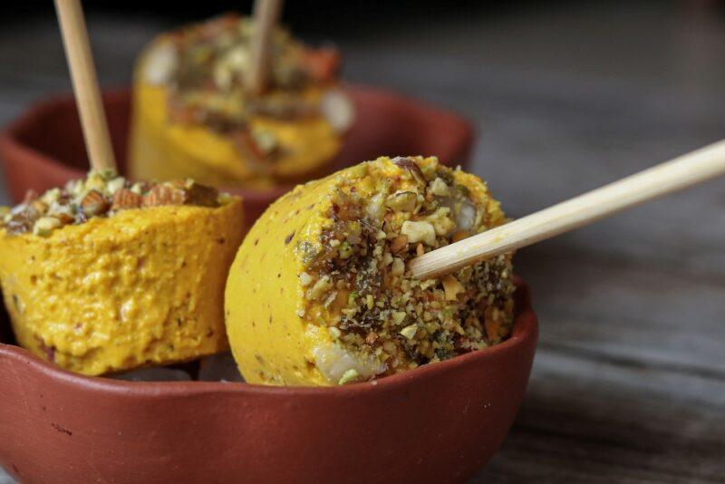 Two brown dishes containing kulfi ice cream on sticks, with spices, seeds, or nuts on the end