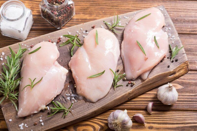 A wooden board with three raw chicken breads and herbs, which will be used to make meat-based noodles