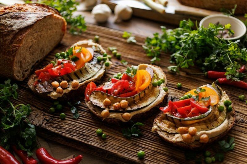 A wooden board with three pieces of sliced sourdough, topped with hummus, tomatoes, grilled zucchini, and a few chickpeas