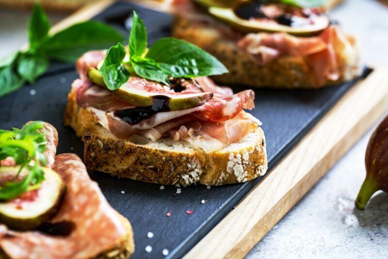 A black slate with three pieces of sourdough that have been topped with sliced prosciutto, figs, and some basil