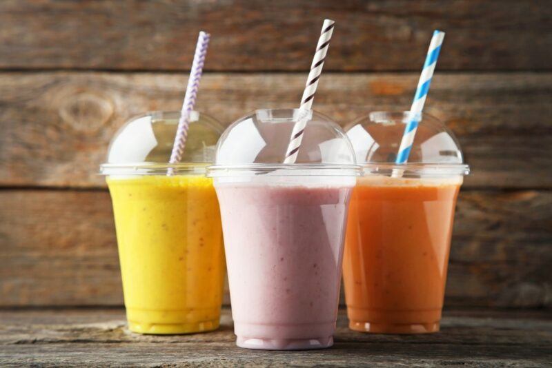 A wooden table with three plastic cups, each containing a different brightly colored smoothie, plus a straw