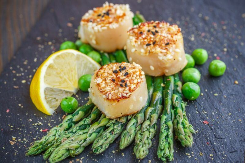 A black table with three cooked scallops, peas, asparagus, and a lemon wedge