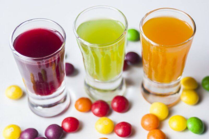 Three shot glasses filled with vodka that's been infused with skittles, there are more skittles across the table