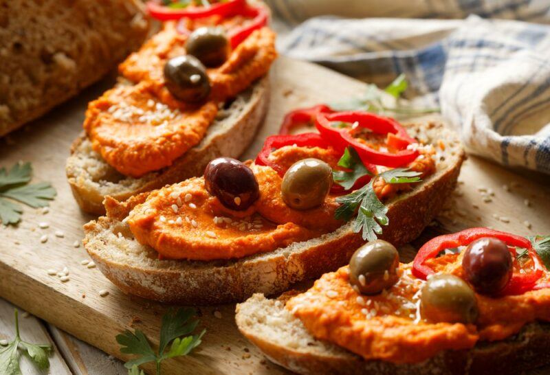 Three slices of sourdough with paprika hummus, fresh olives, and grilled peppers