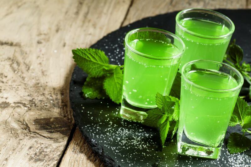 A black tray containing three small glasses of a sour apple Mountain Dew cocktail on a wooden table