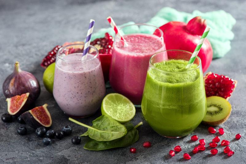 Four fruit smoothies on a table with various types of fruit, like figs, kiwis, and limes