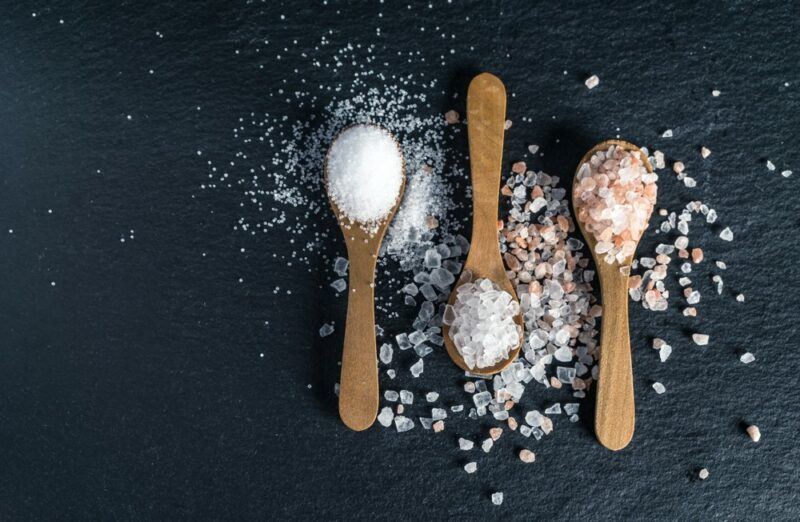 A dark table with three spoons containing different types of salt, including fine table salt, sea salt, and Himalayan salt