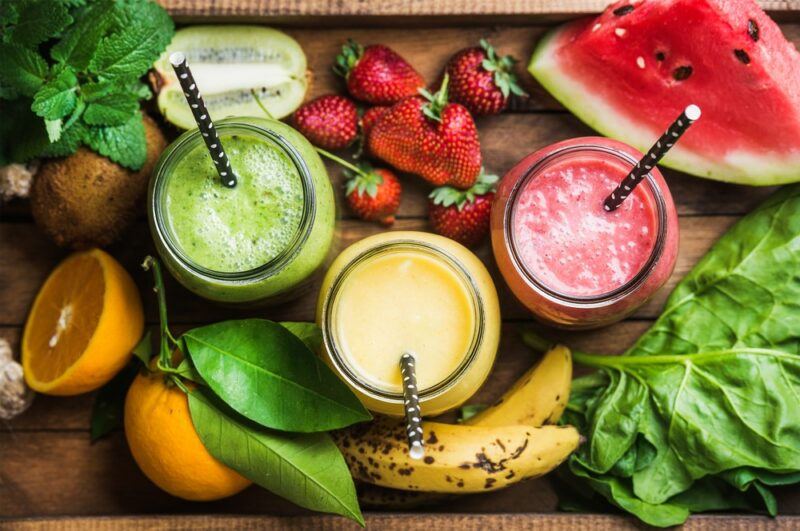 Three mason jars, one with a green smoothie, one pink and one yellow one, along with plenty of fruit