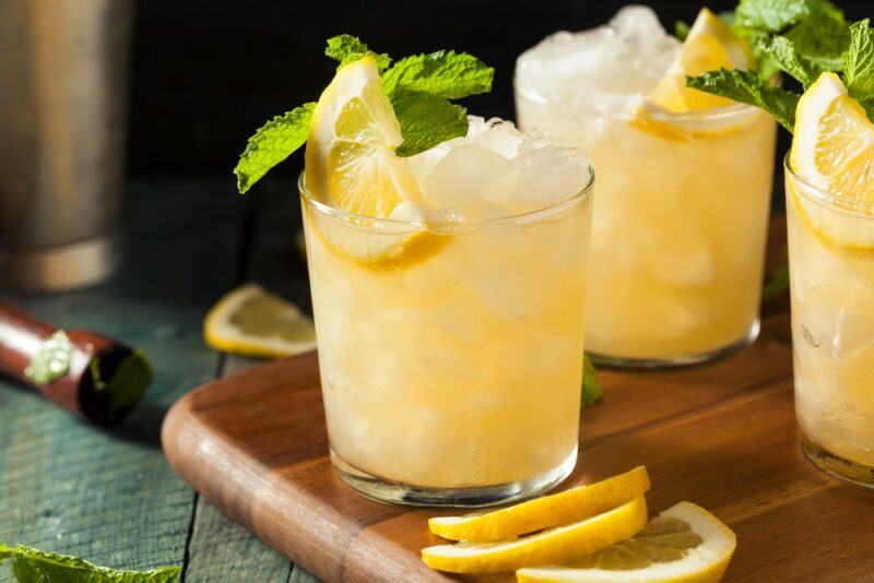 A wooden tray with three glasses of whiskey smash cocktail with a lemon slice and peppermint