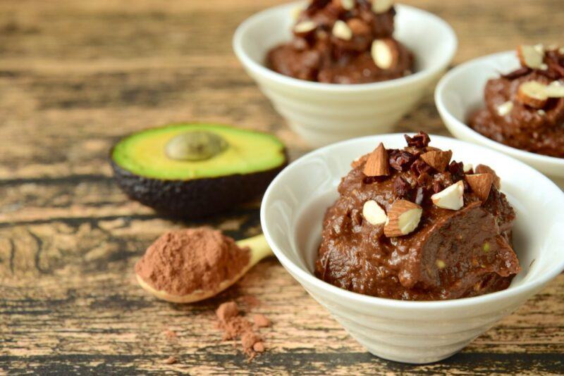 White bowls containing avocado chocolate mousse with nuts, next to half an avocado and some cocoa powder