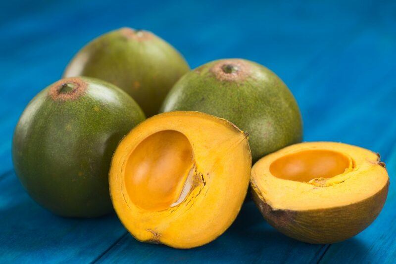 A blue table with three green lucuma fruits, plus two lucuma halves, where you can see the bright orange flesh