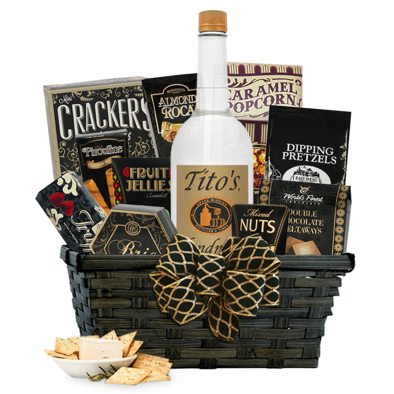 Dark rectangle wicker basket with black and gold bowl with Titos vodka, various snacks including crackers, cheese, pretzels, and nuts. Small bowl with crackers and cheese in front of the left corner of the basket