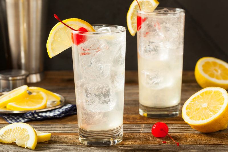 Two glasses of a Tom Collins cocktail with lemons and a glace cheery on a table