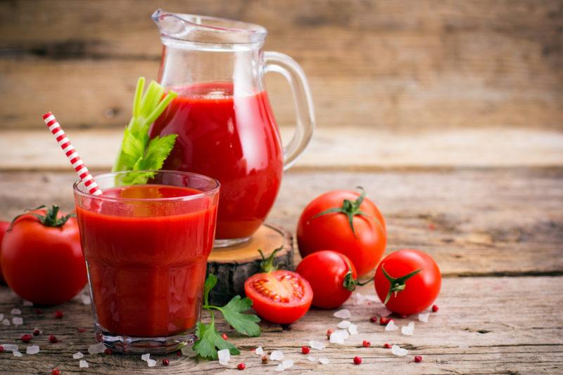 A glass jug of tomato juice, next to a glass of the juice with a straw, some fresh tomatoes, and salt scattered on the table