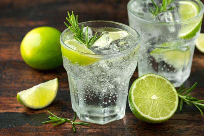 Two glasses containing tonic water, ice and limes, with cut limes on the table