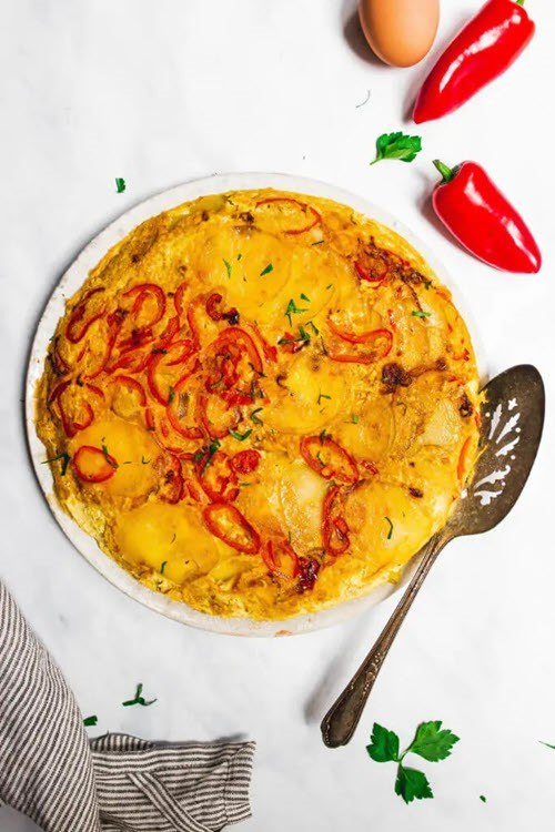 A large white bowl containing a tortilla espanola made with sweet red peppers, with a spoon and peppers on the table