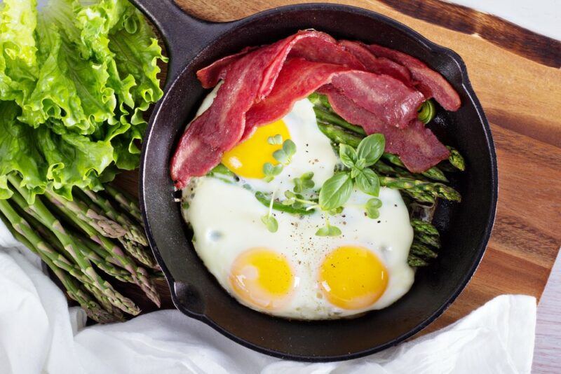 A black frypan with eggs and turkey bacon on a wooden table next to some lettuce